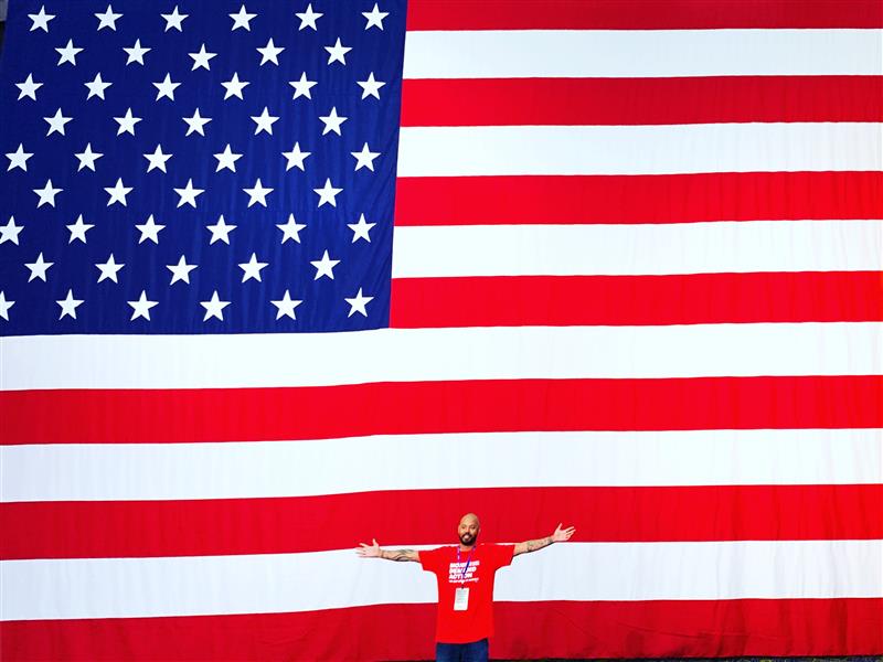 Khary Penebaker posing in front of giant American flag wearing Moms Demand Action shirt