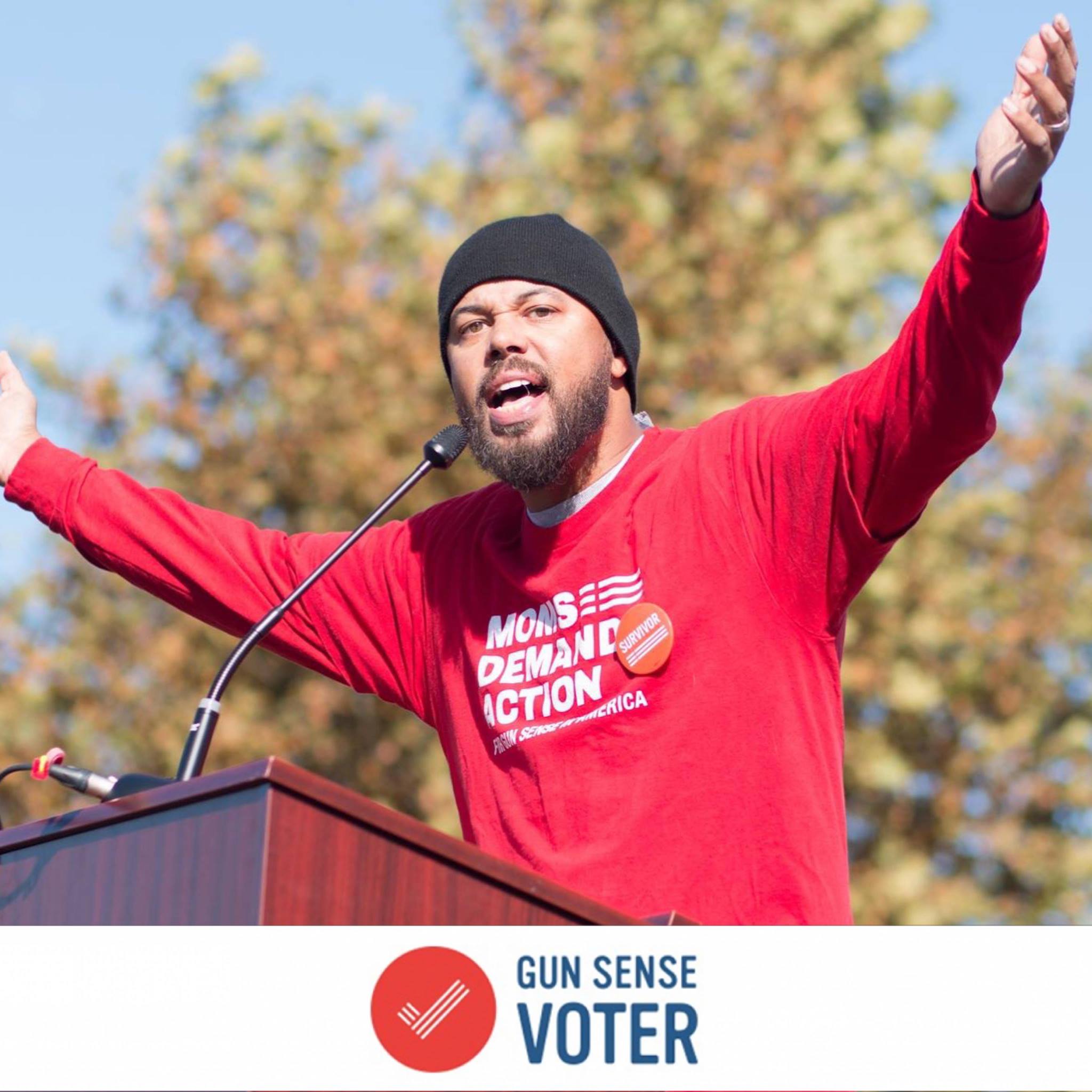 Khary Penebaker at rally podium with arms raised, wearing Moms Demand Action shirt with Gun Sense Voter badge