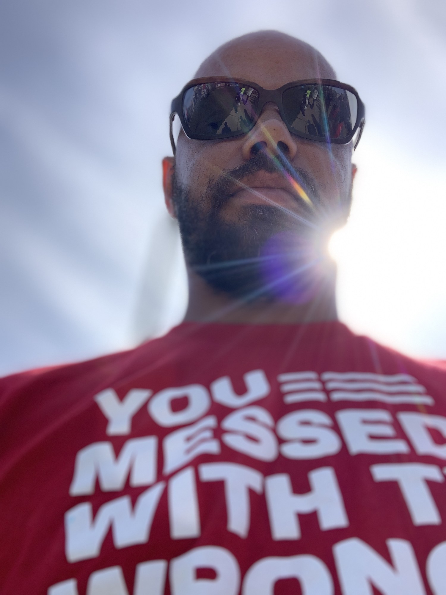 Khary Penebaker portrait at a gun violence prevention rally, low-angle defiant pose with sun flare, wearing a red advocacy t-shirt